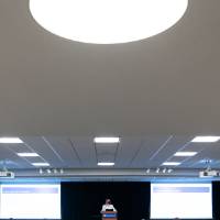 Far shot of audience from behind and scholar wearing mask giving oral presentation.